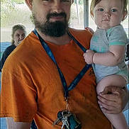 Alex joined the competition — help win amazing prizes! man, baby, child, holding, orange_shirt, cap, beard, keys, lanyard, outdoor, wooden_roof, casual, person, portrait, serious, blue_eyes, short_sleeves, concrete_floor, background_person, natural_light