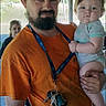 man, baby, child, holding, orange_shirt, cap, beard, keys, lanyard, outdoor, wooden_roof, casual, person, portrait, serious, blue_eyes, short_sleeves, concrete_floor, background_person, natural_light