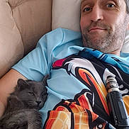 Risko participe au concours pour gagner de l'argent avec cette photo : animal, beige, blue_shirt, cartoon_print, casual, cat, couch, cuddling, facial_hair, gray_cat, human, indoors, man, pet, pillow, relaxed, resting, shirt, smile, vape_device