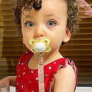 Nelya participe au concours pour gagner de l'argent avec cette photo : baby_accessory, blinds, blue_eyes, child, closeup, curly_hair, cute, expression, face, holding_object, indoor, infant, pacifier, person, portrait, red_dress, skin, toddler, window, young_child