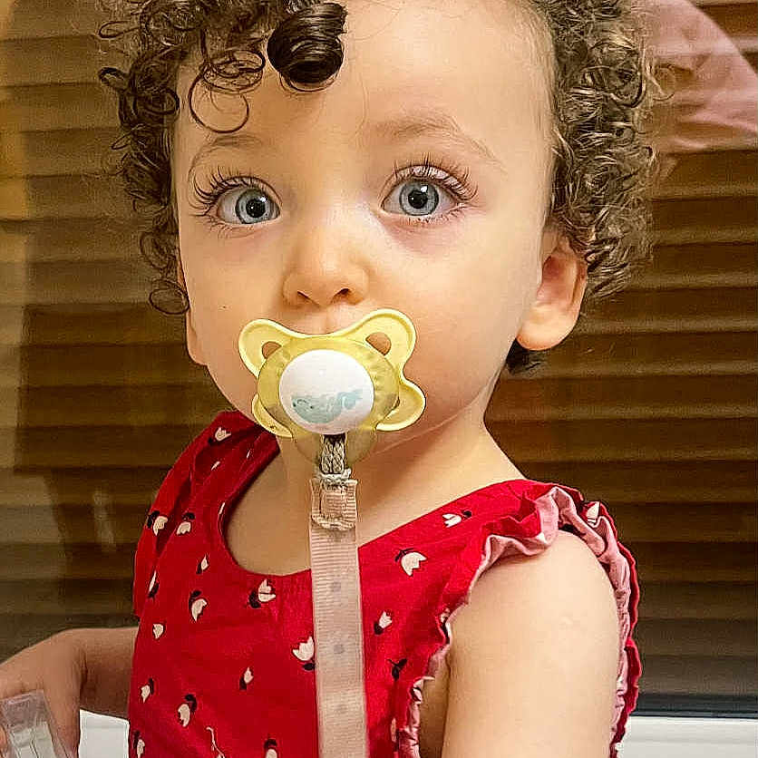 Nelya participe au concours pour gagner de l'argent avec cette photo : baby_accessory, blinds, blue_eyes, child, closeup, curly_hair, cute, expression, face, holding_object, indoor, infant, pacifier, person, portrait, red_dress, skin, toddler, window, young_child