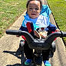 Asaiah is registered to the contest to win money with this photo: child, clothing, cute, daylight, grass, handlebars, happy, jacket, outdoor, parked_cars, person, playtime, residential_area, sidewalk, smiling, socks, sunny, toddler, toy, tricycle