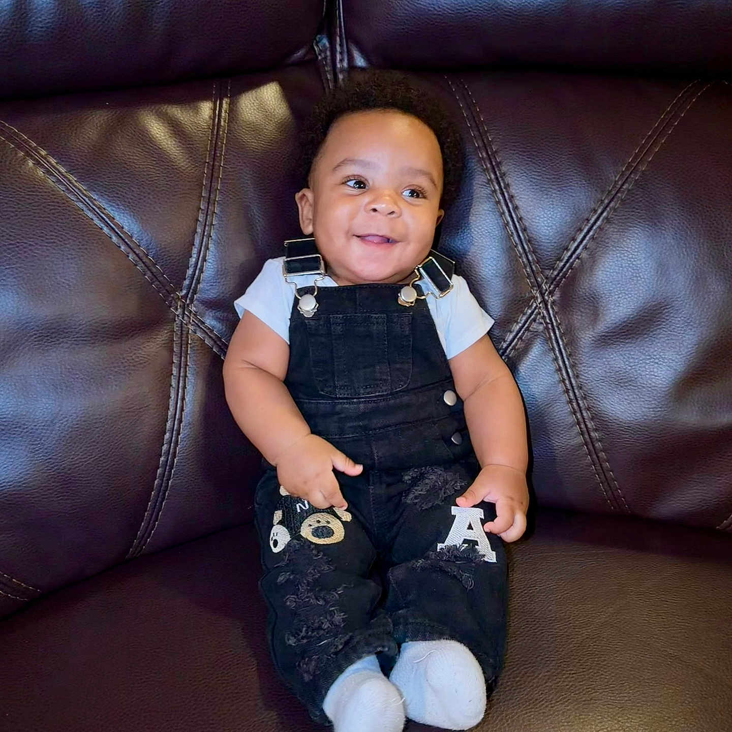 Asaiah joined the competition — help win amazing prizes! baby, child, smiling, overalls, sitting, leather_couch, happy, infant, cute, portrait, clothing, indoor, person, young_child, footwear, black_overalls, white_shirt, baby_socks, seated, joyful