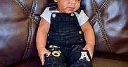 Asaiah is registered to the contest to win money with this photo: toddler, child, baby, overalls, black_clothing, white_shirt, socks, leather_couch, seated, indoor, cute, curious_expression, portrait, young_child, furniture, home, casual_clothing, small_hands, short_hair, relaxed