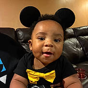 Asaiah is registered to the contest to win money with this photo: baby, child, mickey_mouse, mouse_ears, yellow_bow_tie, black_shirt, smiling, face, indoor, couch, leather, person, cute, happy, toddler, hand, portrait, wall, background, clothing