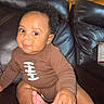 baby, brown_outfit, child, curly_hair, expression, face, foot, football_design, furniture, hand, indoor, infant, leather_couch, light, playful, portrait, room, seated, skin, smile