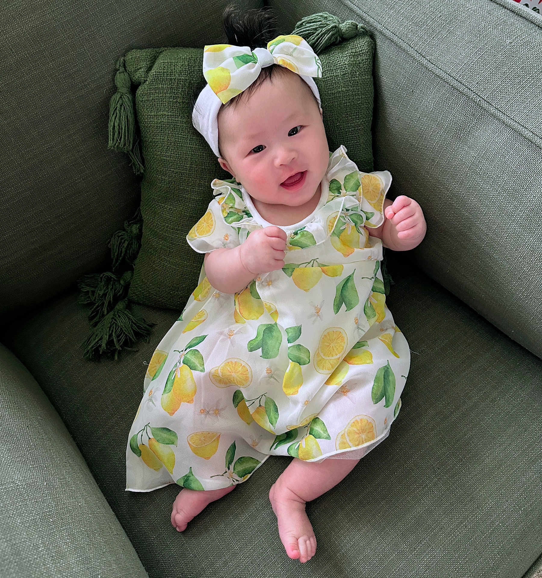 Eliana is registered to the contest to win money with this photo: baby, infant, smiling, lemon_print, dress, headband, bow, couch, green_pillow, tassel, legs, toes, sitting, indoor, portrait, cute, happy, fabric, pattern, person