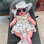 Eliana joined the competition — help win amazing prizes! baby, infant, child, sunglasses, heart_pattern, sun_hat, bouncer, onesie, smile, outfit, legs, feet, indoor, carpet, couch, pillow, wooden_table, accessory, playful, portrait