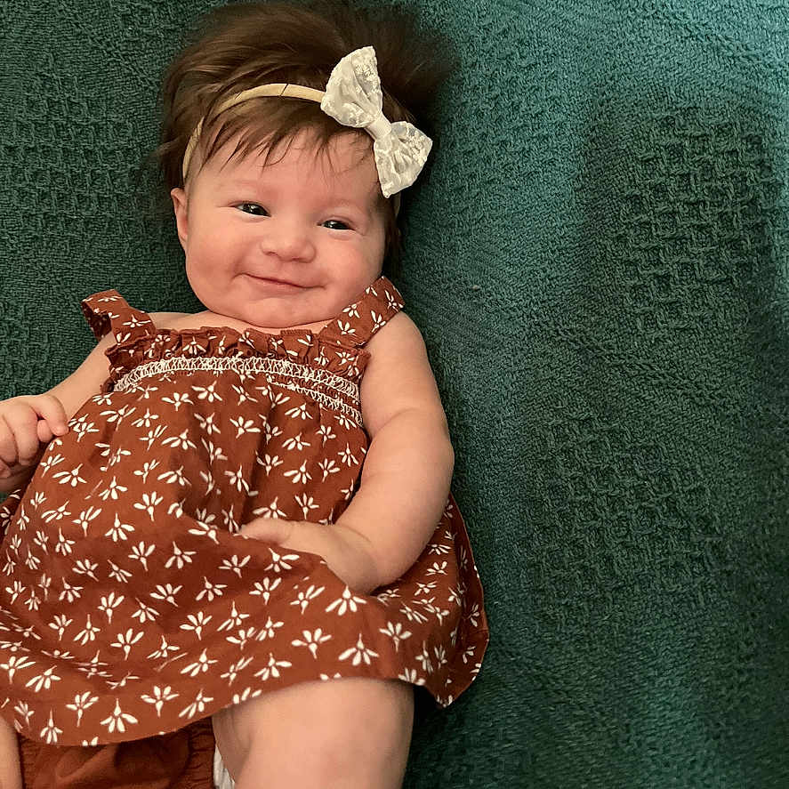 Willow is registered to the contest to win money with this photo: baby, bodypart, bonnet, cap, clothing, couch, dress, face, finger, furniture, hand, happy, hat, head, newborn, person, photography, portrait, sitting, smile