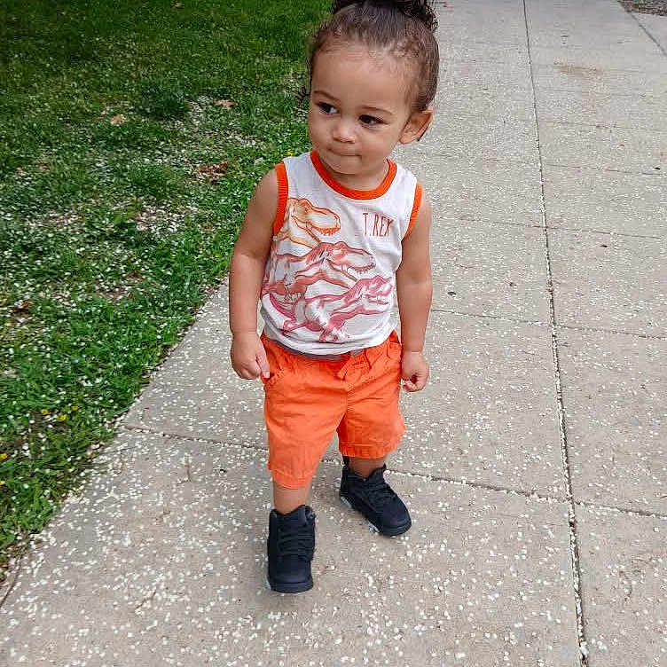 Marquis is registered to the contest to win money with this photo: toddler, child, sidewalk, grass, orange_shorts, tank_top, t_rex, curly_hair, black_shoes, outdoor, street, urban, casual_clothing, summer, young_child, portrait, walking, cute, daytime, person