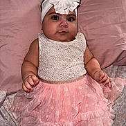 Kiara is registered to the contest to win money with this photo: baby, child, infant, portrait, sitting, bed, pink, headband, flower, dress, skirt, polka_dots, ruffles, wide_eyes, face, hands, indoors, bedding, cute, fashion