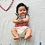 baby, infant, smiling, lying_down, diaper, striped_clothing, black_hair, hand_clasp, pink_beads, heart_shape, white_blanket, cute, chubby_cheeks, portrait, child, happy, soft_lighting, indoors, close_up, person