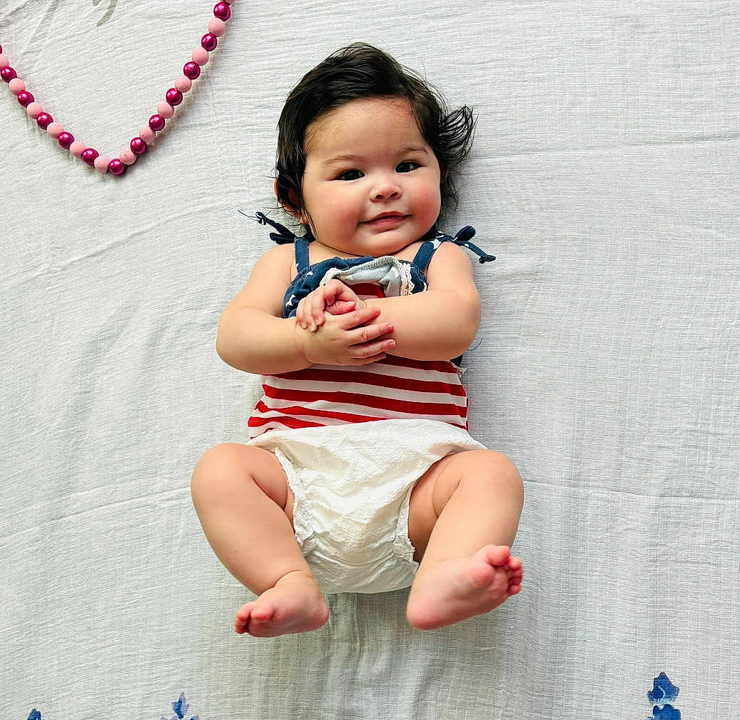 Scarlett is registered to the contest to win money with this photo: baby, infant, smiling, lying_down, diaper, striped_clothing, black_hair, hand_clasp, pink_beads, heart_shape, white_blanket, cute, chubby_cheeks, portrait, child, happy, soft_lighting, indoors, close_up, person