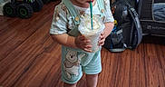 Elias is registered to the contest to win money with this photo: toddler, child, indoor, wooden_floor, toy_trucks, overalls, bear_print, sandal, drink, cup, straw, curly_hair, playpen, bag, door, floor_transition, person, home, happy, casual_clothing