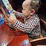 toddler, child, menu, restaurant, high_chair, wooden_table, curly_hair, checkered_shirt, indoor, focused, seated, table, food_menu, baby, person, side_view, casual_clothing, young_child, curiosity, furniture