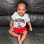 baby, casual_clothing, child, comfortable, cute, face, feet, hand, happy, indoors, infant, leather_couch, person, portrait, shorts, sitting, skin, smiling, tshirt, young