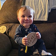 George joined the competition — help win amazing prizes! baby, child, couch, blanket, wooden_sign, onesie, socks, smile, indoor, furniture, face, person, happy, seated, home, cozy, infant, holding, toy, window