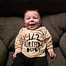 baby, smiling, infant, child, cute, sitting, clothing, shirt, text, happy, indoors, couch, furniture, portrait, young_child, face, hands, casual, relaxed, cozy