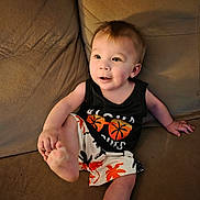 George is registered to the contest to win money with this photo: toddler, child, smiling, couch, clothing, tank_top, shorts, indoor, face, person, foot, happy, sitting, palm_tree, patterned_clothing, cute, infant, casual, relaxed, young_child