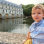 child, toddler, boy, striped_shirt, blue_bandana, stone_railing, river, bridge, historic_building, water, boats, trees, outdoor, daylight, portrait, casual_clothing, nature, calm, reflection, travel