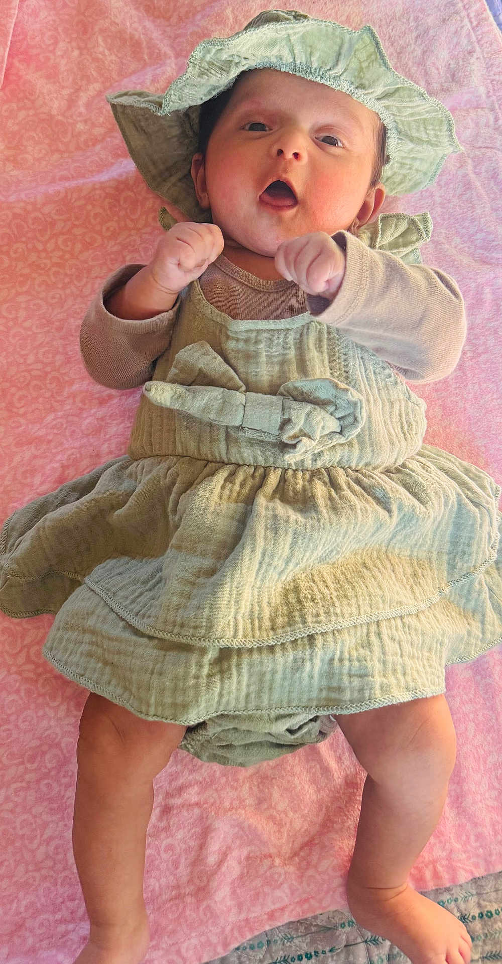 Sanvika joined the competition — help win amazing prizes! baby, infant, hat, dress, blanket, pink_background, hands, legs, feet, cute, portrait, lying_down, bow, fabric, skin, cheeks, surprised_expression, green_clothing, bed, newborn