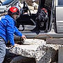 dog, black_dog, person, helmet, blue_jacket, vehicle, car, open_door, concrete, rocks, outdoor, rescue, training, leash, rubble, workwear, safety_gear, daylight, action, caution