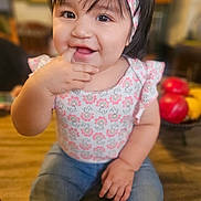 Eva is registered to the contest to win money with this photo: baby, child, smiling, headband, floral, cute, indoor, jeans, sitting, happy, portrait, fingers_in_mouth, table, fruit, red_apple, yellow_fruit, blurred_background, person, clothing, young