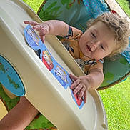 Chevy is registered to the contest to win money with this photo: toddler, child, high_chair, outdoor, grass, cards, curly_hair, playing, baby, greenery, happy, seat, toy, person, nature, sunlight, summer, colorful, smiling, hands