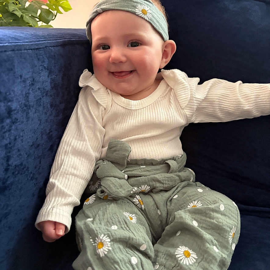 Ivy is registered to the contest to win money with this photo: baby, child, smiling, headband, green_pants, daisy_print, white_top, couch, navy_blue, indoor, plant, curtains, happy, sitting, portrait, cute, infant, home, cozy, clothing