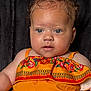 Zaaryah is registered to the contest to win money with this photo: baby, child, face, blue_eyes, curly_hair, orange_dress, floral_pattern, portrait, cute, infant, closeup, skin, person, young_child, fashion, colorful_clothing, expression, sitting, indoors, background