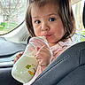 child, toddler, girl, baby_bottle, drinking, pink_clothing, hair_bows, brown_hair, portrait, face, eyes, indoors, car, vehicle, passenger, seated, hands, milk, cute, daylight