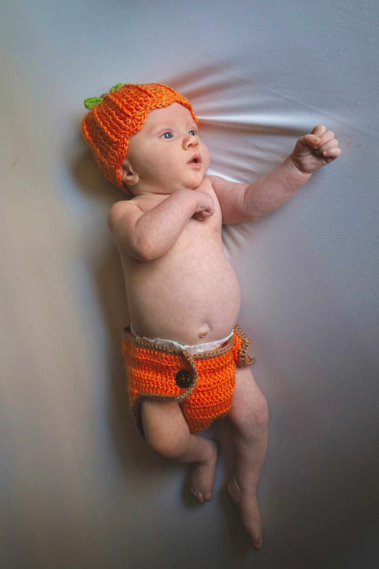 Elijah joined the competition — help win amazing prizes! baby, infant, knitted_hat, orange, diaper_cover, lying_down, curious, skin, arm, leg, cute, child, newborn, expression, background, soft_surface, photography, portrait, clothing, button