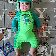 Gabriel joined the competition — help win amazing prizes! baby, child, dinosaur_outfit, green_clothing, play_mat, animal_illustrations, colorful, infant, cute, smiling, lying_down, indoors, headwear, hat, face, feet, hands, bright_colors, pattern, soft_surface