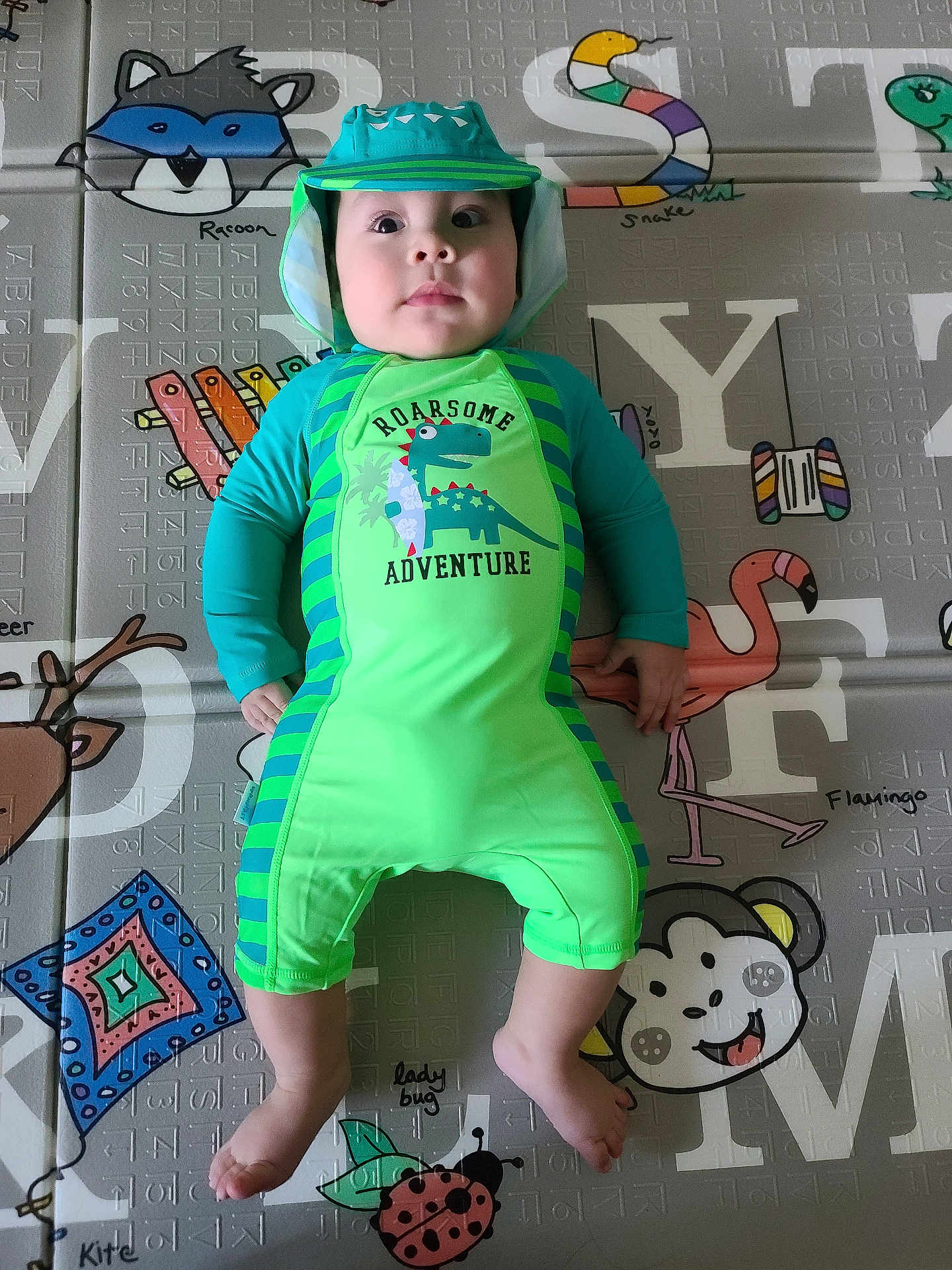 Gabriel joined the competition — help win amazing prizes! baby, child, dinosaur_outfit, green_clothing, play_mat, animal_illustrations, colorful, infant, cute, smiling, lying_down, indoors, headwear, hat, face, feet, hands, bright_colors, pattern, soft_surface