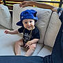 baby, child, couch, cap, blue_cap, gold_chain, smile, sitting, indoor, person, arm, black_tshirt, beige_cushion, window, daylight, wooden_blinds, cute, happy, portrait, casual