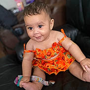 Kenleanna joined the competition — help win amazing prizes! baby, child, smiling, orange_dress, sitting, black_couch, indoor, happy, cute, infant, person, portrait, clothing, skin, hand, foot, colorful, patterned, young, seat