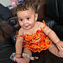 baby, child, smiling, orange_dress, sitting, black_couch, indoor, happy, cute, infant, person, portrait, clothing, skin, hand, foot, colorful, patterned, young, seat