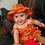 Kenleanna is registered to the contest to win money with this photo: baby, child, orange_hat, orange_dress, floral_pattern, smiling, happy, plush_toy, red_toy, yellow_accents, sitting, indoor, leather_chair, cute, portrait, infant, playful, person, clothing, hat