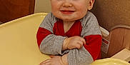 Reilly is registered to the contest to win money with this photo: baby, child, high_chair, smiling, indoor, person, red_clothing, gray_clothing, wooden_chair, floor, happy, face, head, infant, sitting, hands, table, cute, young, portrait