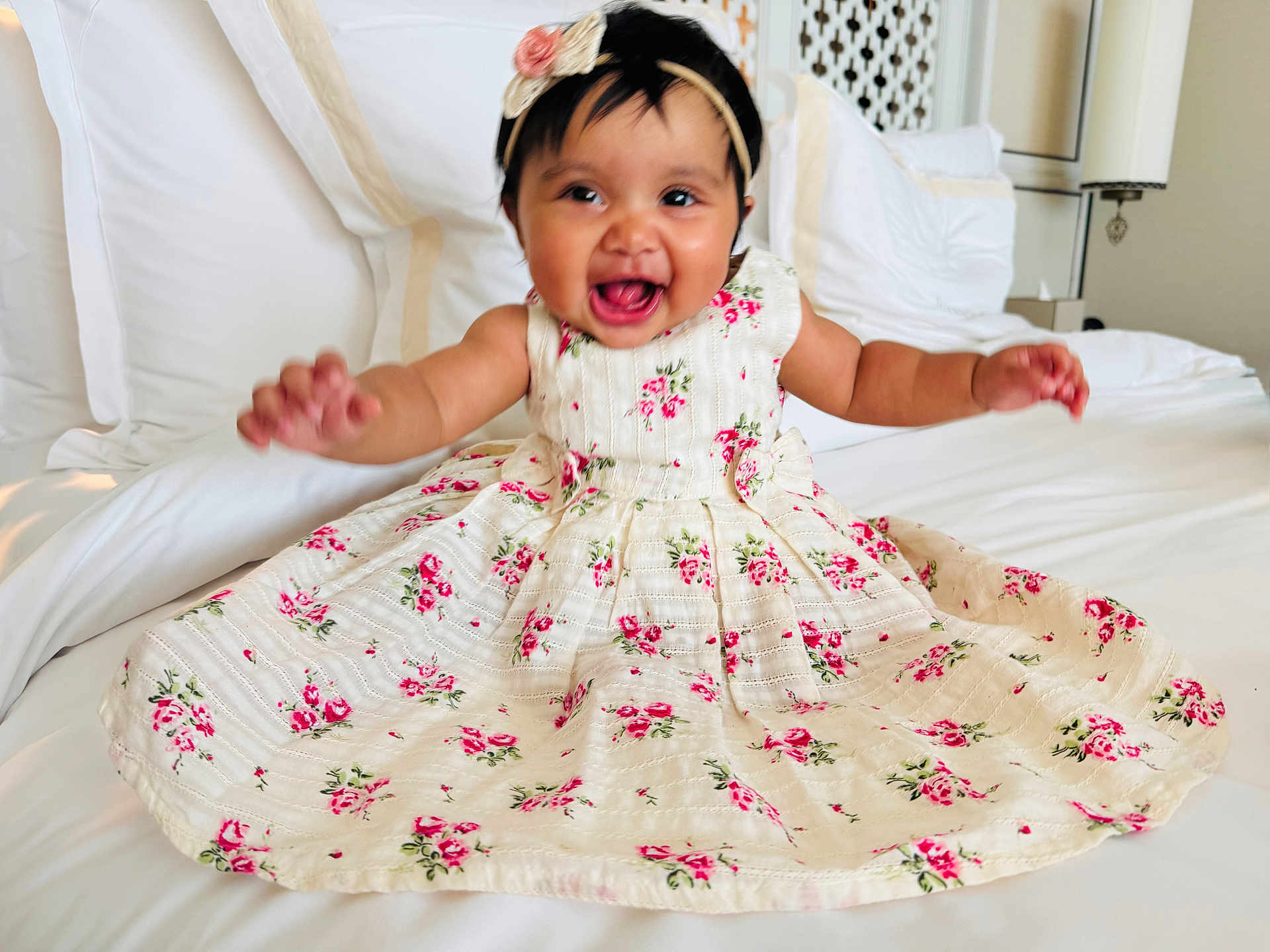 Liyana is registered to the contest to win money with this photo: baby, child, smiling, dress, floral_pattern, headband, flower, bed, white_bed, happy, cute, indoors, person, portrait, sitting, young_child, cheerful, fabric, clothing, skin