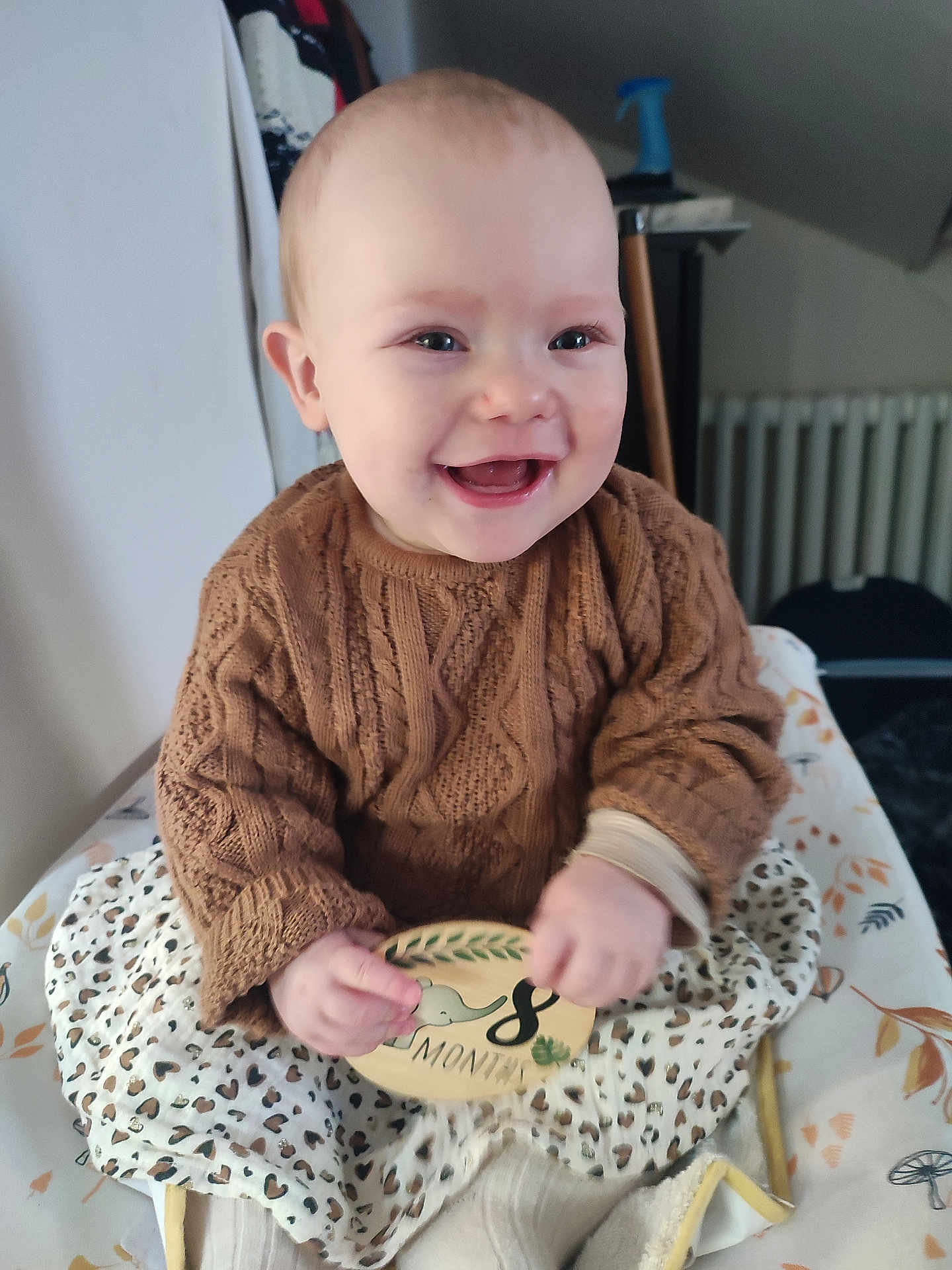 Elyna participe au concours pour gagner de l'argent avec cette photo : baby, child, smiling, knitted_sweater, brown_sweater, plaque, 8_months, patterned_skirt, blanket, indoor, happy, portrait, sitting, cozy, cute, infant, clothing, hands, face, young_child