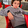 child, person, red_chair, gray_sweater, headband, indoor, cafe, table, casual_clothing, modern_interior, relaxed_pose, young, face, seated, watch, black_jeans, lighting, blurred_background, portrait, confident