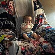 Oliver is registered to the contest to win money with this photo: toddler, child, phone, chair, blanket, family_photos, indoor, portrait, casual_clothing, shorts, shirt, barefoot, light, wall, framed_photos, curious, seated, home, cozy, soft_light