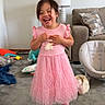child, girl, pink_dress, smiling, laughing, living_room, carpet, lamp, sofa, pillow, toy, shoes, happy, standing, indoor, cute, playful, home, young_child, toddler