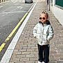 child, kid, puffer_jacket, silver_jacket, sunglasses, sidewalk, cobblestone, street, urban, casual, standing, shoes, cool, confident, fashion, outdoor, daytime, person, clothing, young