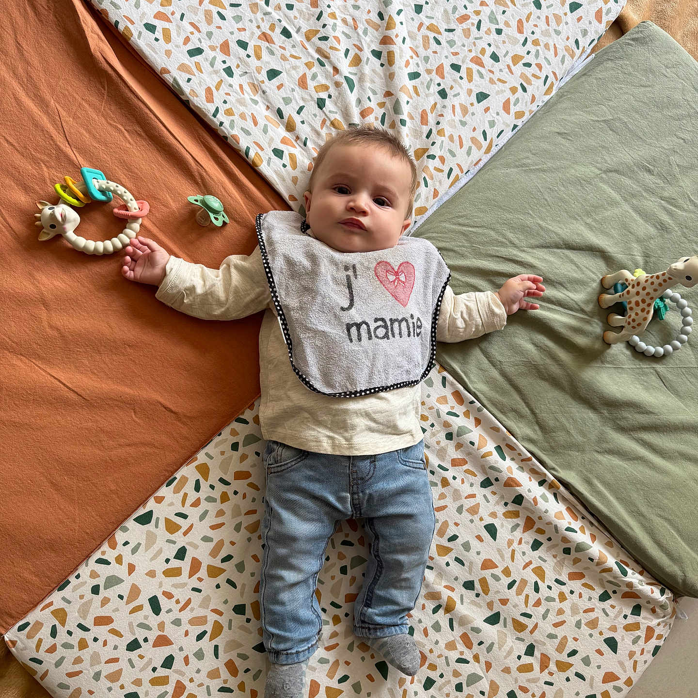 Maëlo participe au concours pour gagner de l'argent avec cette photo : baby, bib, child, clothing, colorful, cute, expression, floor, indoor, infant, jeans, laying_down, pacifier, pattern, person, playtime, portrait, quilt, socks, toy
