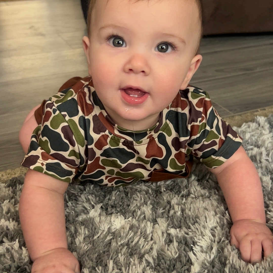 Reece joined the competition — help win amazing prizes! baby, child, crawling, carpet, floor, camouflage_shirt, drool, indoor, living_room, curious, hands, face, eyes, person, cute, toddler, plush, home, furniture, wood_floor