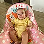 Liam is registered to the contest to win money with this photo: baby, infant, child, yellow_onesie, baby_seat, pink, polka_dots, animal_print, flooring, wooden_floor, indoor, kitchen, appliances, cute, relaxed, sitting, barefoot, person, portrait, home