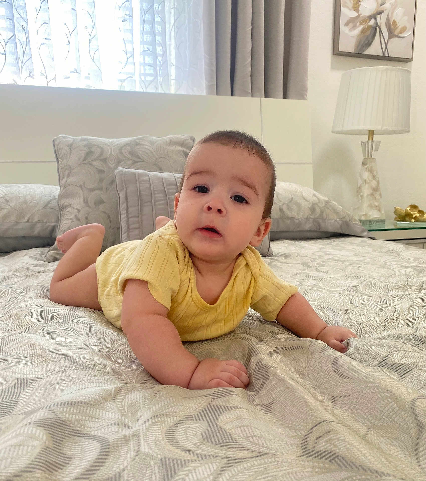 Liam is registered to the contest to win money with this photo: baby, infant, bed, yellow_clothing, pillow, blanket, curtain, lamp, bedroom, person, face, expression, indoors, furniture, soft_light, head, hands, pattern, resting, toddler