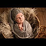 baby, basket, child, cozy, cute, expression, fur, headwear, indoor, infant, knit_hat, looking, newborn, person, portrait, seated, soft_texture, swaddle, wooden_floor, wrapped
